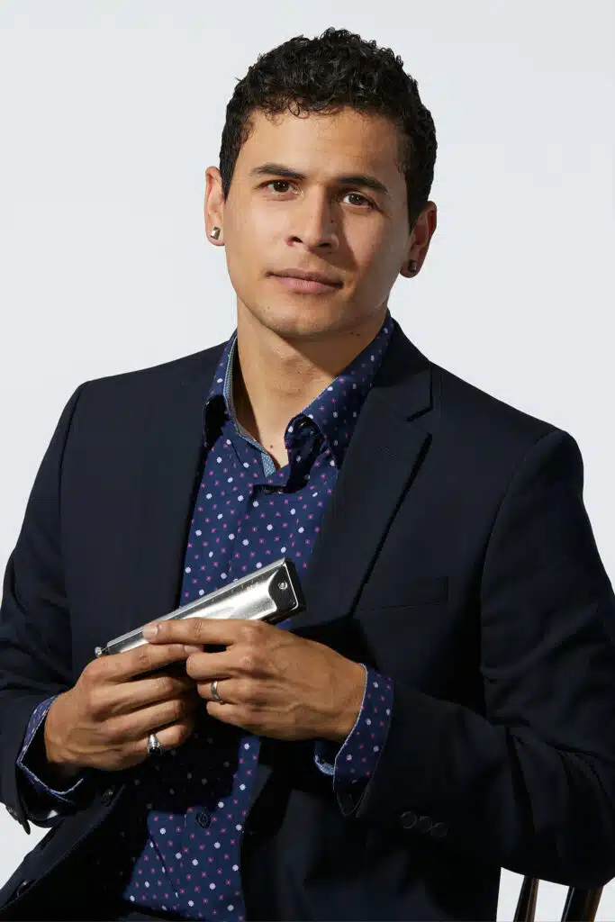 Alan posing in front of camera wearing a blue button up with polka dotted design and a black blazer holding a harmonica